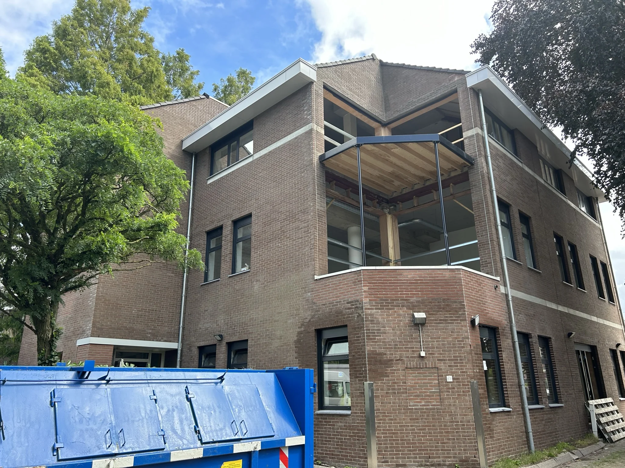 Bakstenen kantoorpand aan de Bergse Dorpsstraat met een opengewerkte hoekverdieping en een blauwe afvalcontainer op de voorgrond.